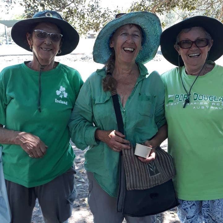Three laughing, smiling women, standing arm in arm, dressed in green and wearing broad brimmed hats. They look like they are good friends.